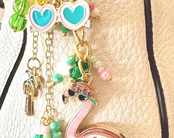 Könnte beinhalten: Ein goldfarbener Schlüsselanhänger mit tropischen Anhängern, darunter ein rosa Flamingo, herzförmige Sonnenbrillen und eine Palme. Die Anhänger sind mit rosa, grünen und transparenten Perlen verziert, was ein lebendiges Accessoire ergibt.