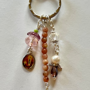 May include: A silver key chain with a round ring and a lobster clasp. The chain has a variety of charms, including a small round image, a cross, a pearl, and several crystals.