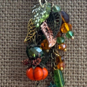 May include: A fall-themed charm with a lobster clasp. It features an orange pumpkin, green and amber beads, and metal leaf charms in bronze and teal. The charm is designed with a variety of textures and colors, perfect for autumn.