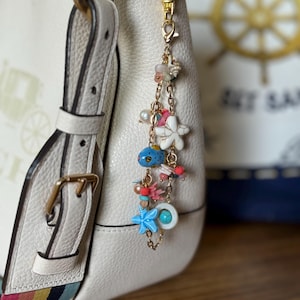 K&ouml;nnte beinhalten: Eine cremefarbene Lederhandtasche mit farbenfrohem Riemen ist mit einem goldfarbenen Anh&auml;nger verziert. Der Anh&auml;nger zeigt Meeresmotive, darunter ein blauer Fisch, Seesterne und Perlen, was eine nautische &Auml;sthetik erzeugt.