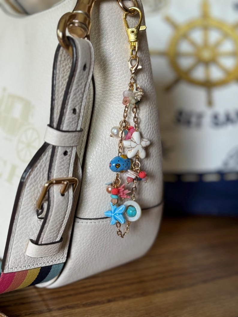 K&ouml;nnte beinhalten: Eine cremefarbene Lederhandtasche mit farbenfrohem Riemen ist mit einem goldfarbenen Anh&auml;nger verziert. Der Anh&auml;nger zeigt Meeresmotive, darunter ein blauer Fisch, Seesterne und Perlen, was eine nautische &Auml;sthetik erzeugt.