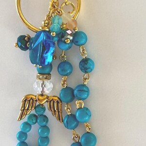 May include: A gold-toned keychain with turquoise beads, a blue crystal, and a gold angel wing charm. Additional charms include a blue ribbon, a pearl, and a gold cross. The keychain is designed with a lobster claw clasp.