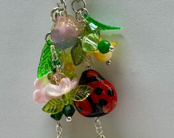 Könnte beinhalten: Ein dekorativer Schlüsselanhänger mit verschiedenen Anhängern. Er zeigt einen roten Marienkäfer mit schwarzen Punkten, grüne Blätter, rosa Blüten und einen Schmetterling. Der Schlüsselanhänger ist an einem silberfarbenen Verschluss befestigt.
