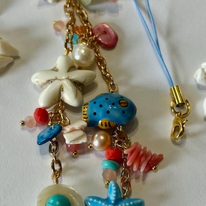 K&ouml;nnte beinhalten: Ein goldfarbenes Charm-Armband mit verschiedenen maritimen Anh&auml;ngern. Die Anh&auml;nger umfassen einen blauen Kugelfisch, wei&szlig;e und blaue Seesterne, Muscheln und bunte Perlen. Ein blaues Band mit einem goldenen Verschluss ist ebenfalls befestigt.