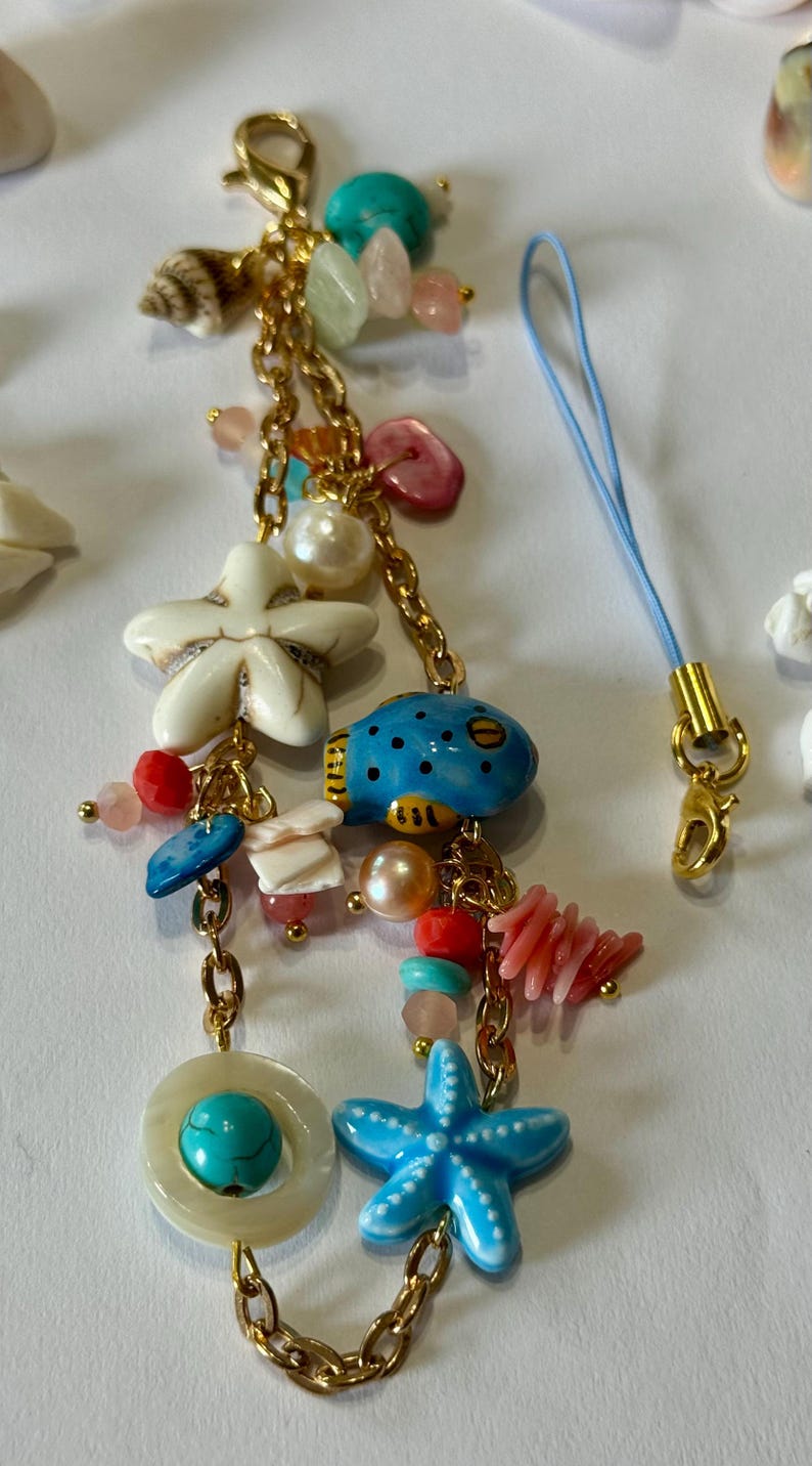 K&ouml;nnte beinhalten: Ein goldfarbenes Charm-Armband mit verschiedenen maritimen Anh&auml;ngern. Die Anh&auml;nger umfassen einen blauen Kugelfisch, wei&szlig;e und blaue Seesterne, Muscheln und bunte Perlen. Ein blaues Band mit einem goldenen Verschluss ist ebenfalls befestigt.