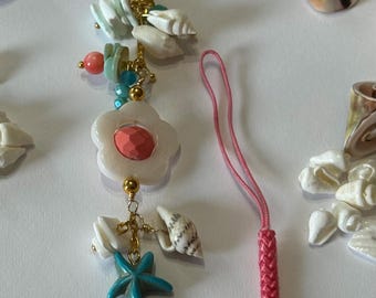 May include: A decorative charm with seashells, turquoise starfish, coral beads, and a white flower pendant. The charm is attached to a gold-coloured clasp and a pink braided lanyard. Various seashells are scattered in the background.