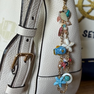 K&ouml;nnte beinhalten: Ein goldfarbener Charm mit verschiedenen Meeresmotiven, darunter ein blauer Seestern, ein blauer Fisch und eine wei&szlig;e Blume. Der Charm ist an einer wei&szlig;en Lederhandtasche mit einer goldenen Schnalle befestigt.