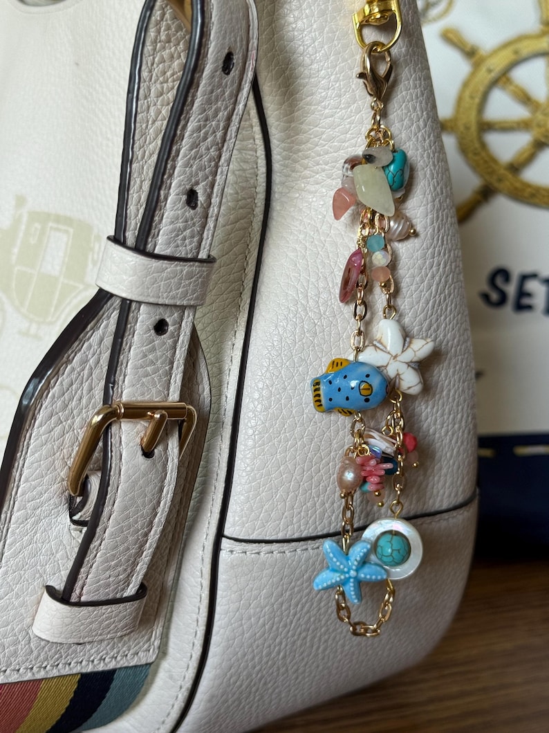 K&ouml;nnte beinhalten: Ein goldfarbener Charm mit verschiedenen Meeresmotiven, darunter ein blauer Seestern, ein blauer Fisch und eine wei&szlig;e Blume. Der Charm ist an einer wei&szlig;en Lederhandtasche mit einer goldenen Schnalle befestigt.
