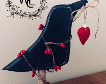 May include: A black wooden bird sculpture with a red felt heart ornament hanging from a twine bow. The bird is decorated with red berry branches and sits on a wooden base. The text "WE Creations" is in a floral wreath in the background.