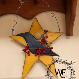 May include: A rustic, yellow star-shaped wall hanging with a distressed finish. A dark blue bird silhouette is centered on the star, adorned with red berries and feathers. The piece is suspended by a wire and features the text "WE est. front. creation's."