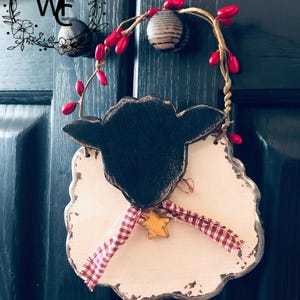May include: A wooden sheep-shaped door hanger with a black head and white body. It features a red and white checkered ribbon, a gold star, and a decorative berry branch. The door hanger is designed with a rustic, farmhouse aesthetic.