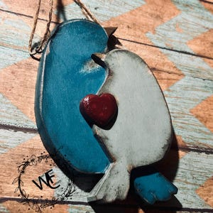 May include: A hanging wooden ornament featuring two stylized birds, one in deep teal and one in light gray, facing each other. A small, dark red heart is positioned between the birds. The ornament includes a jute twine loop for hanging.
