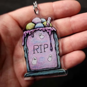 May include: A pastel pink and purple tombstone with a white RIP inscription and three white ghosts. The tombstone is topped with a pink and white swirl of frosting and a straw. The tombstone is a keychain.