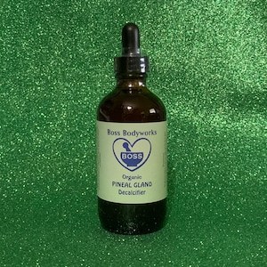 Op de afbeelding: Een bruine glazen fles met een druppelaar, met het label "Boss Bodyworks" en "Organic Pineal Gland Decalcifier". Het label heeft een hart met een vijzel en stamper erin, en het woord "Boss" in blauw.