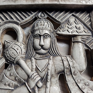 Könnte beinhalten: Eine weiße Keramik-Reliefskulptur einer Gottheit mit einer Krone, die einen Stab und einen belaubten Zweig hält. Die Skulptur ist mit aufwendigen Schnitzereien verziert und hat eine strukturierte Oberfläche.