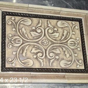 May include: Rectangular decorative tile with a beige and brown color scheme. The center features a floral design with swirling leaves and a small flower. The tile is framed with a rope-like border and measures 17 3/4 x 23 1/2 inches.