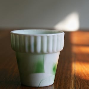 May include: A small white ceramic planter with green and white marbling. The planter has a fluted rim and is sitting on a wooden surface.