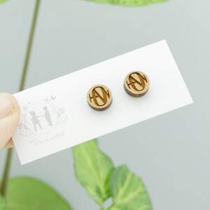 May include: A pair of wooden stud earrings with a circular design. Each earring features a carved letter "N" within a circle. The earrings are displayed on a white card with the text "ADAM & EVE" and a small illustration.