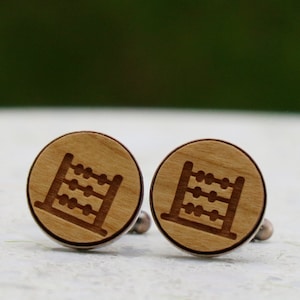 Könnte beinhalten: Ein Paar runde Manschettenknöpfe aus Holz mit einem gelaserten Abakus-Design. Die Manschettenknöpfe haben eine hellbraune Holzmaserung und eine silberfarbene Metallrückseite. Das Abakus-Design ist dunkelbraun.