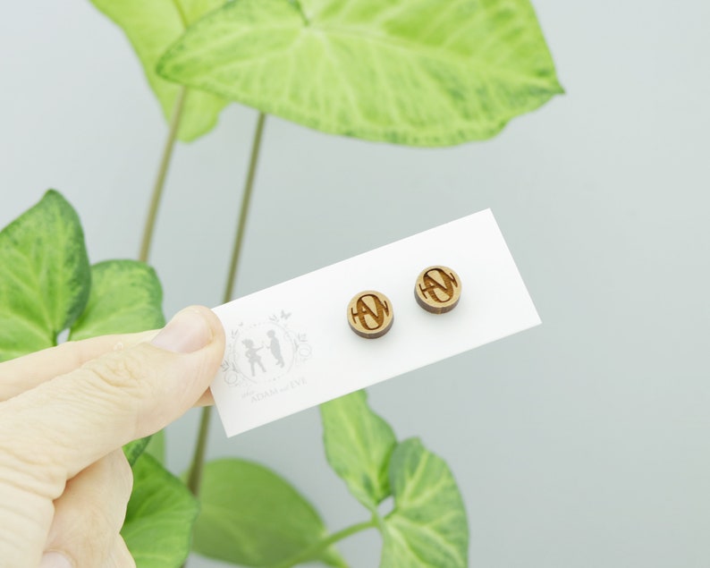 May include: A pair of wooden stud earrings with a circular design. Each earring features a carved letter "N" within a circle. The earrings are displayed on a white card with the text "ADAM & EVE" and a small illustration.