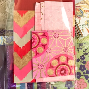 May include: A collection of pink and gold patterned paper scraps with geometric and floral designs. The paper is arranged in a layered stack, with the top layer featuring a pink background with gold geometric shapes and floral accents.