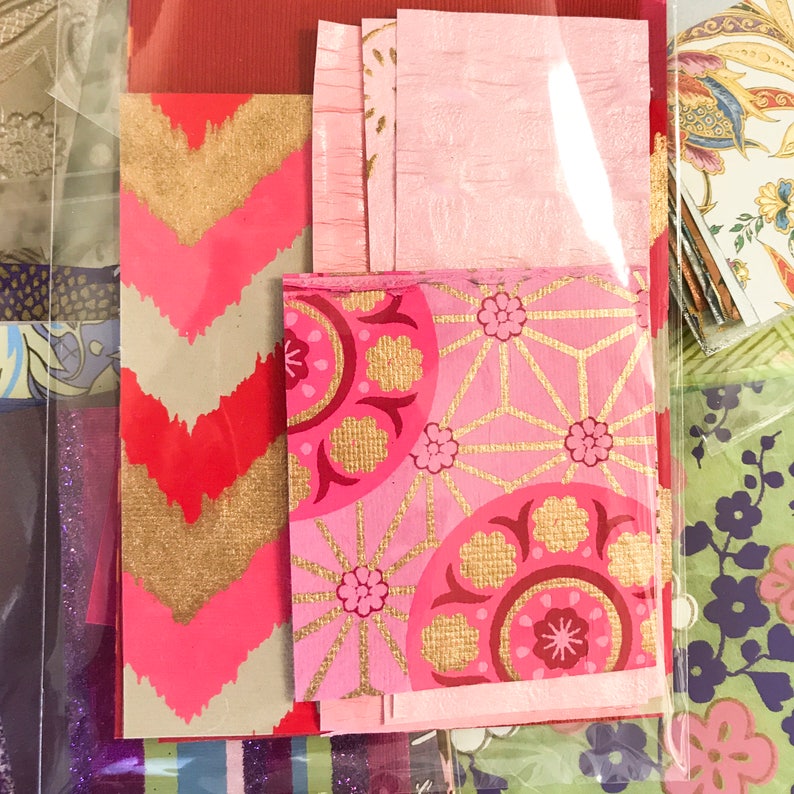May include: A collection of pink and gold patterned paper scraps with geometric and floral designs. The paper is arranged in a layered stack, with the top layer featuring a pink background with gold geometric shapes and floral accents.