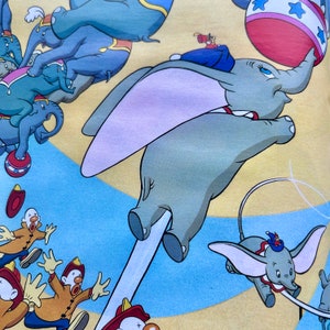 May include: A colorful illustration of Dumbo the elephant flying with his ears. He is a light gray color with pink ears and a blue and white striped tail. There are other elephants in the background, as well as a red and white striped ball.