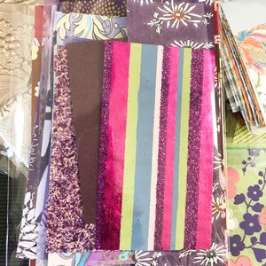 May include: A collection of patterned paper scraps in various shades of purple, pink, and green. The scraps are arranged in a vertical stack, with a striped piece of paper with pink, green, and purple glitter on top.