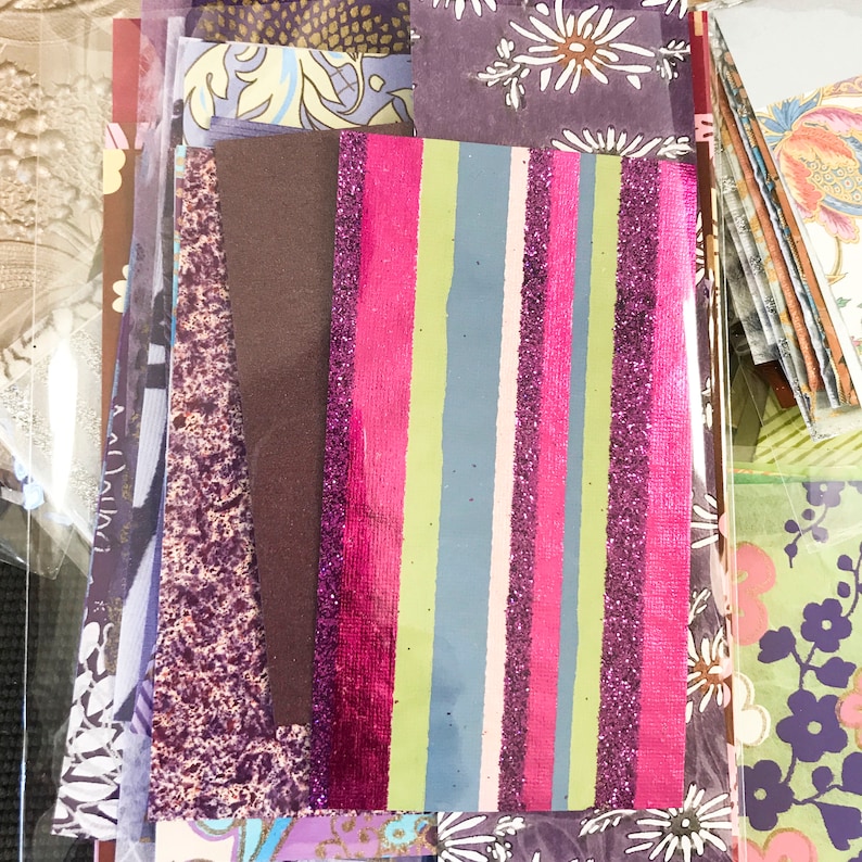 May include: A collection of patterned paper scraps in various shades of purple, pink, and green. The scraps are arranged in a vertical stack, with a striped piece of paper with pink, green, and purple glitter on top.