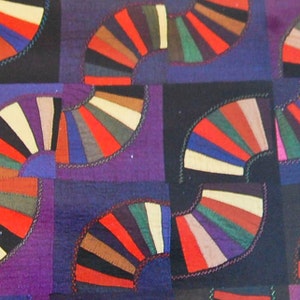 May include: A quilt with a repeating pattern of colorful fan-shaped designs. The quilt is made of various fabrics in shades of red, blue, green, brown, and white. The background is a deep purple color.