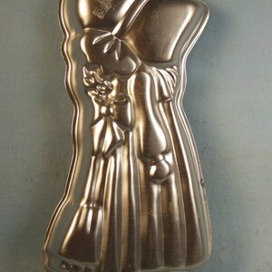May include: A silver metal cake pan shaped like a girl wearing a bonnet and a dress. The pan has a textured surface and is designed for baking cakes.