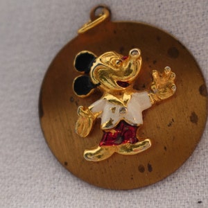 Op de afbeelding: Een gouden metalen hanger met een cartoon Mickey Mouse met zwarte oren, een wit shirt, een rode broek en een gouden omtrek. De hanger is rond en heeft een lus aan de bovenkant om aan een ketting of armband te bevestigen.