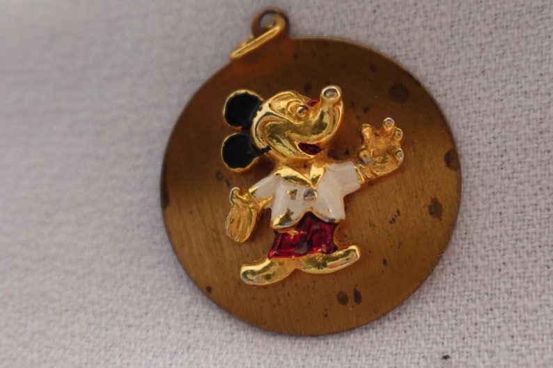 Op de afbeelding: Een gouden metalen hanger met een cartoon Mickey Mouse met zwarte oren, een wit shirt, een rode broek en een gouden omtrek. De hanger is rond en heeft een lus aan de bovenkant om aan een ketting of armband te bevestigen.