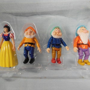 May include: A set of five plastic figurines from the Disney movie Snow White and the Seven Dwarfs. The set includes Snow White, who is wearing a yellow dress with a red and blue bodice, and four dwarfs. The dwarfs are wearing colorful clothing and have white beards.
