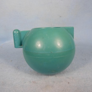 May include: A teal-colored, spherical plastic container with a handle on the side. The container has a circular opening at the top and a horizontal seam around the middle. The handle is rectangular and extends outward.