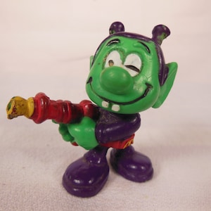 May include: A green alien toy figure with purple boots and a red and yellow ray gun. The alien has a winking eye and a toothy grin.