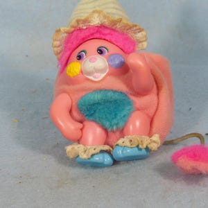 May include: A vintage pink plush toy with a blue tuft, blue shoes, and a hat with a pink brim. The toy has a white face, yellow and purple accents, and a pink tail with a fluffy end. The toy is a collectible item.