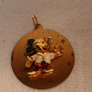Op de afbeelding: Een gouden hanger met een cartoon Mickey Mouse in een wit shirt met een rode broek en zwarte schoenen. De hanger is rond en heeft een getextureerd oppervlak.