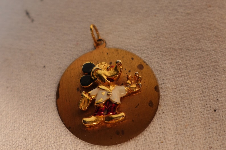 Op de afbeelding: Een gouden hanger met een cartoon Mickey Mouse in een wit shirt met een rode broek en zwarte schoenen. De hanger is rond en heeft een getextureerd oppervlak.