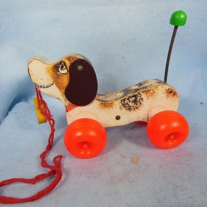 Op de afbeelding: Vintage houten trekdier speelgoed in de vorm van een hond met oranje wielen en een rood trekkoord. De hond heeft een bruin oor, een zwart oog en een groene knop bovenop. Het speelgoed is beschilderd met een gevlekt patroon.