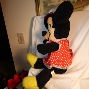 May include: A large plush toy of Minnie Mouse wearing a red dress with white polka dots and a white lace trim. The toy has black fur and large black ears with a red bow.