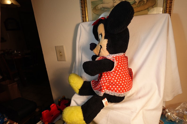 May include: A large plush toy of Minnie Mouse wearing a red dress with white polka dots and a white lace trim. The toy has black fur and large black ears with a red bow.