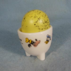 Puede incluir: Un huevo amarillo moteado en una huevera de cerámica blanca. La huevera tiene tres patas pequeñas y está decorada con gráficos de personajes de dibujos animados. El fondo es azul claro liso.