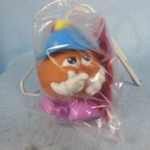 Puede incluir: Un juguete de Mr. Potato Head, empaquetado en una bolsa de plástico transparente. El juguete tiene una cabeza marrón, un sombrero azul, guantes blancos y zapatos morados. El juguete tiene una nariz roja y un acento amarillo en el sombrero.