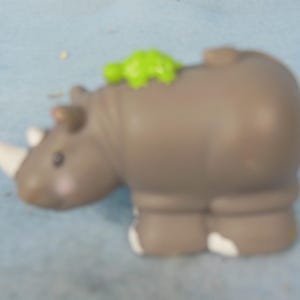 May include: A gray toy rhinoceros with a white horn and a small green figure on its back. The toy has white accents on its feet and is set against a light blue background. This toy is designed for children.