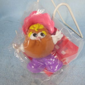 Puede incluir: Un juguete nuevo y sin abrir de Mr. Potato Head en una bolsa de plástico transparente. El juguete es marrón con un sombrero rosa, pelo amarillo y una falda morada. El juguete está empaquetado con una etiqueta roja y un asa blanca.