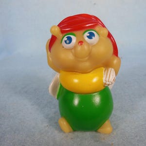 Könnte beinhalten: Vintage Kunststoff-Spielfigur einer cartoonartigen, insektenartigen Figur. Die Figur hat einen beigefarbenen Körper, rotes Haar, blaue Augen und ein gelbes Hemd. Der untere Körper ist grün, und sie hat kleine, beige Füße.