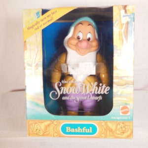Puede incluir: Una muñeca de plástico de Tímido, uno de los siete enanitos de Blancanieves y los Siete Enanitos de Disney. La muñeca lleva una chaqueta amarilla, una camisa blanca, pantalones morados y un sombrero azul. La cara de la muñeca está pintada con un rubor rosado y tiene una expresión de sorpresa. La muñeca está empaquetada en una caja con las palabras "Blancanieves y los Siete Enanitos" y "Tímido" impresas en ella.