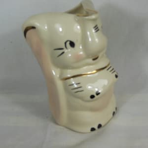 May include: A ceramic pitcher in the shape of an elephant, with a cream-colored body, pink accents, and black and gold details. The elephant has a smiling face and is decorated with black lines and dots.