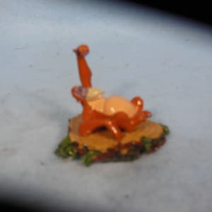 Puede incluir: Una pequeña figura naranja de un personaje de dibujos animados sentado en un tocón de árbol marrón. El personaje tiene la barriga rosa y sostiene un pequeño objeto morado.